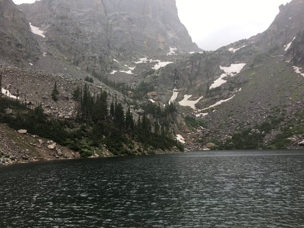 Alpine lake surrounded by rocky cliffs, forested slopes, and patches of snow, viewed from the shoreline in a mountain… — Rocky Mountain National Park