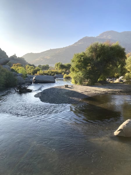 A flowing river with boulder-strewn banks and riparian vegetation, viewed from the water's edge with mountains rising… — Sequoia & Kings Canyon