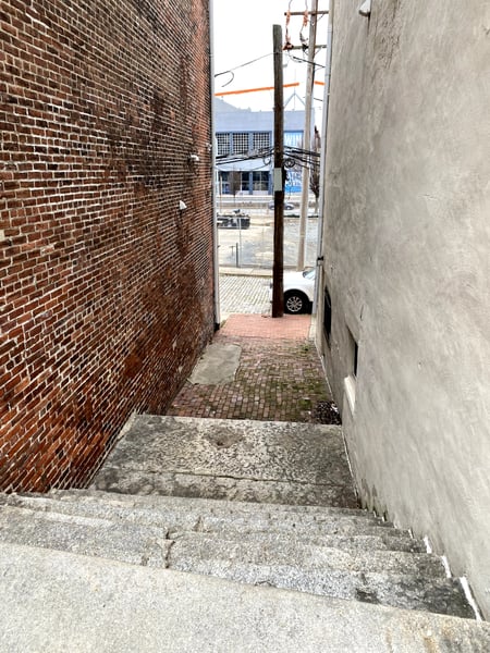 Narrow brick alleyway between industrial buildings viewed from street level, opening to a waterfront street in… — Philadelphia