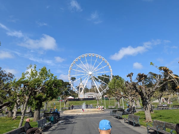 A white Ferris wheel rises above Rideout Fountain in a public park, framed by mature trees and green lawns on a clear… — Inner Sunset, San Francisco