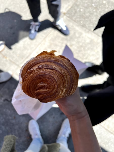 A chocolate croissant pastry held up by hand on a sunny street with pedestrians visible in the background. — Inner Richmond, San Francisco