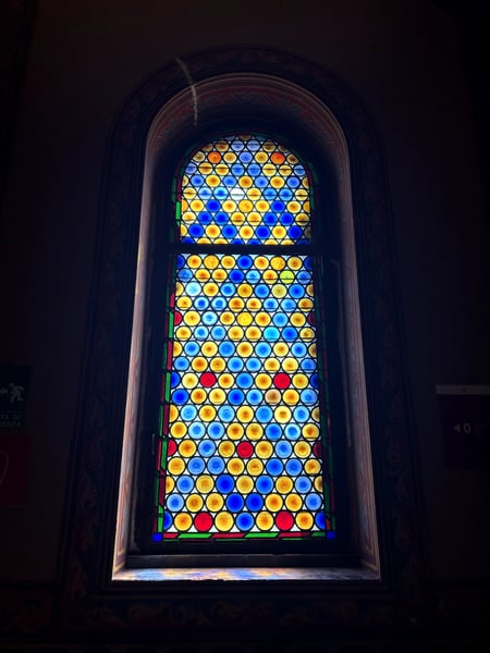Modern stained glass window with blue, yellow, and red circular patterns in a dark stone archway, viewed from inside a… — Oltrarno, Florence