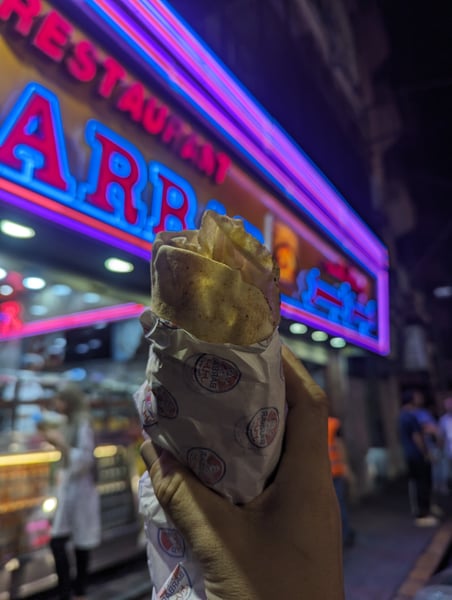 Street food being held up against neon-lit storefront signage at night in an urban setting. — Beirut
