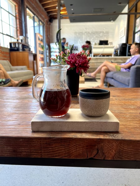 A pitcher of iced tea and ceramic cup on a wooden table in a modern café or hotel lobby with red flowers and guests in… — Los Angeles