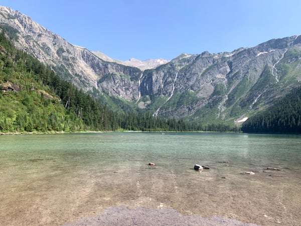 Turquoise alpine lake surrounded by forested mountain peaks and waterfalls, viewed from the shoreline. — Glacier National Park