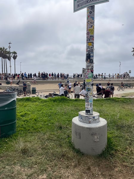 Sticker-covered utility pole on Venice Beach grass with beachgoers and cyclists in the background. — Venice, Los Angeles