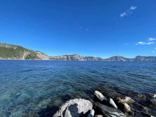 Clear blue lake waters with rocky shoreline and forested mountain cliffs visible across the water at Cleetwood Cove. — Crater Lake National Park