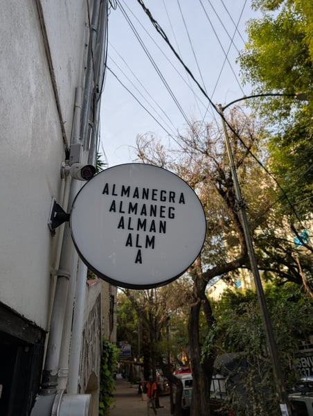 Circular cafe sign for Almanegra reading vertically, mounted on a white building wall in a Mexico City street with…