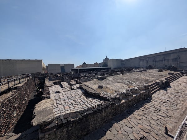 Ancient Roman archaeological ruins with stone foundations and mosaic floors viewed from ground level, with modern city… — Centro, Mexico City