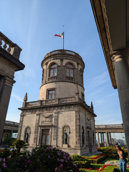 Neoclassical stone tower with circular gallery and Mexican flag, framed by colonnaded portico and manicured gardens. — San Miguel Chapultepec, Mexico City