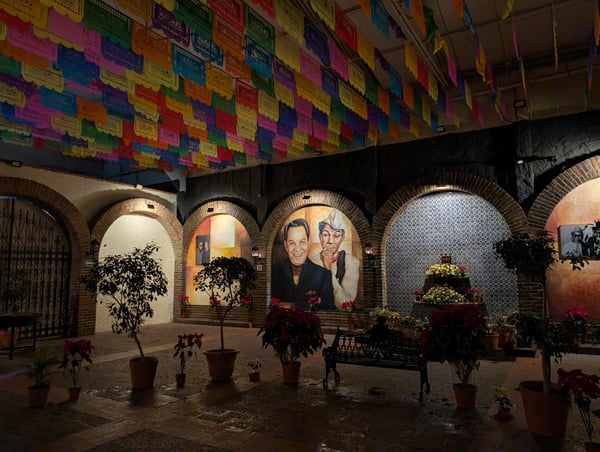 Colorful papel picado decorations hanging above a brick courtyard with portrait murals and potted flowers. — Mexico City