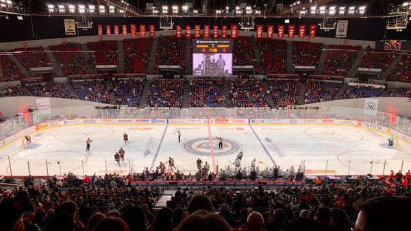Ice hockey game in progress at a packed arena with red seating, viewed from the crowd level. — Portland