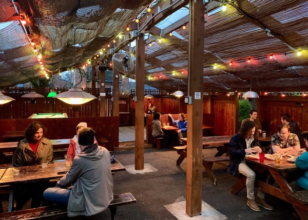 Paymaster Lounge's covered outdoor beer garden with wooden booths, string lights, and patrons dining at dusk in… — Northwest District, Portland