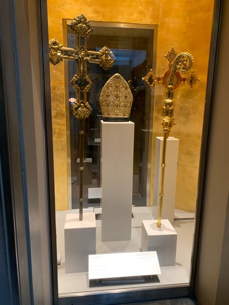Golden ornamental ecclesiastical objects including crosses and a bishop's mitre displayed in a museum case with ochre… — Piazza del Duomo, Florence