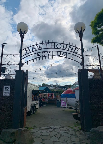 Hawthorne Asylum entrance gate with decorative arch, string lights, and food vendors visible beyond in Portland, Oregon.