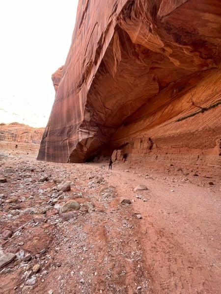 A hiker stands beneath a massive red sandstone overhang in a desert canyon landscape. — The Wave Trail, Kanab & The Wave