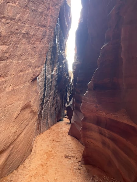 Narrow red rock slot canyon with towering sandstone walls and bright sky visible through the opening, taken from ground… — The Wave Trail, Kanab & The Wave