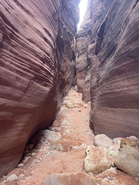 Narrow slot canyon with towering red rock walls and sandy floor, viewed from within the passage looking toward daylight. — The Wave Trail, Kanab & The Wave