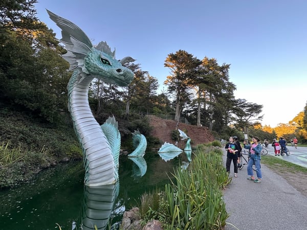 A large turquoise dragon sculpture in a park pond with visitors on a walking path nearby during autumn. — San Francisco