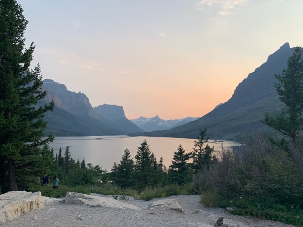 Alpine lake surrounded by coniferous forest and steep mountain peaks at sunset, viewed from a rocky overlook. — Glacier National Park
