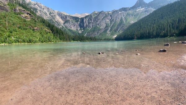 Turquoise alpine lake surrounded by coniferous forests and rocky mountain peaks with waterfalls, viewed from a sandy… — Glacier National Park