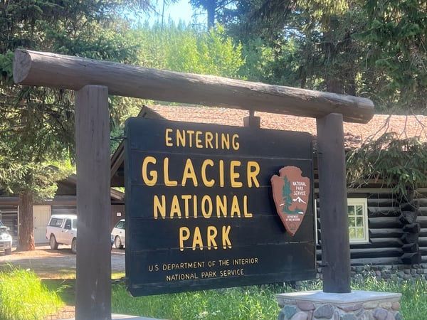 Entrance sign to Glacier National Park with National Park Service emblem, surrounded by coniferous forest.